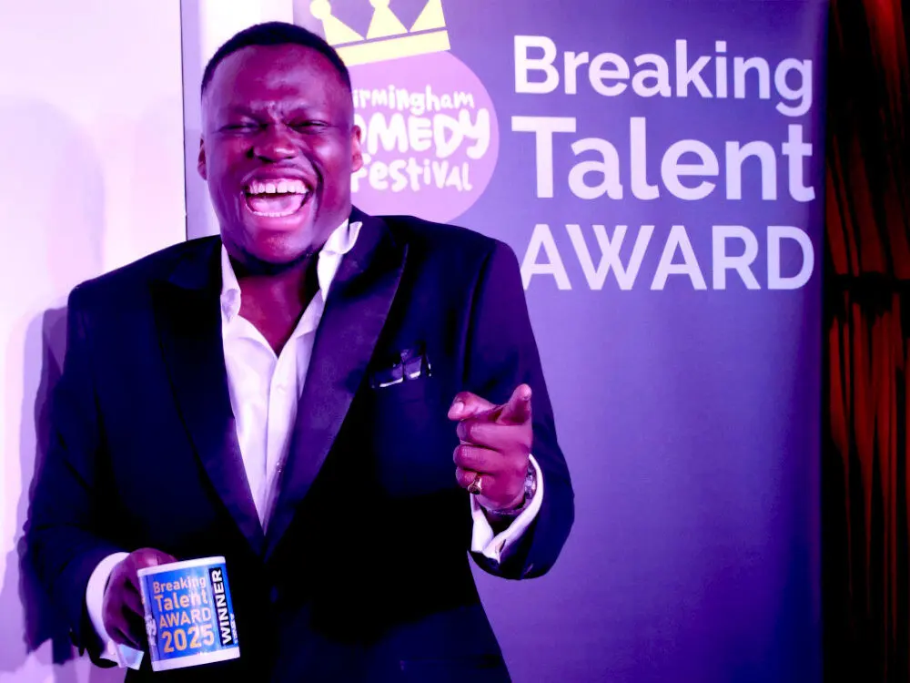 The 2025 Breaking Talent Award winner laughing on stage with his winner's mug in front of the Birmingham Comedy Festival