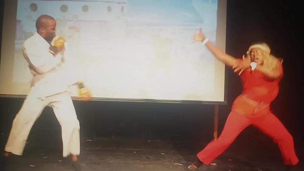 Two performers in a high-energy sketch on stage in front of a projection screen at the Black Women in Comedy Festival