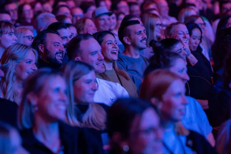 A smiling, laughing audience in purple light at the festival's 1LIVE Comedy-Nacht XXL in the LANXESS arena.