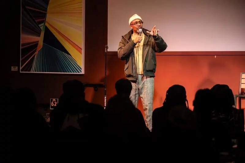 A comedian on a small club stage with the audience in silhouette at the DC Comedy Festival