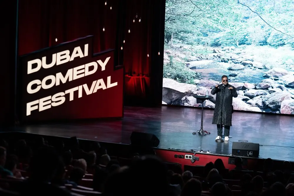 A comedian performing on a big theatre stage beside the festival's own red stage sign, with the audience in the foreground