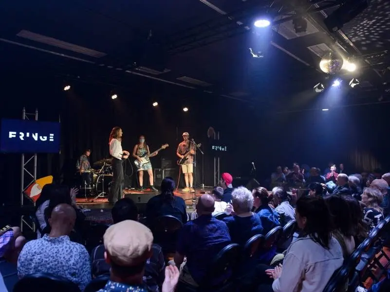 A band on a lit stage at the Dunedin Fringe 2023 opening party, the Fringe sign at left and a crowd watching.