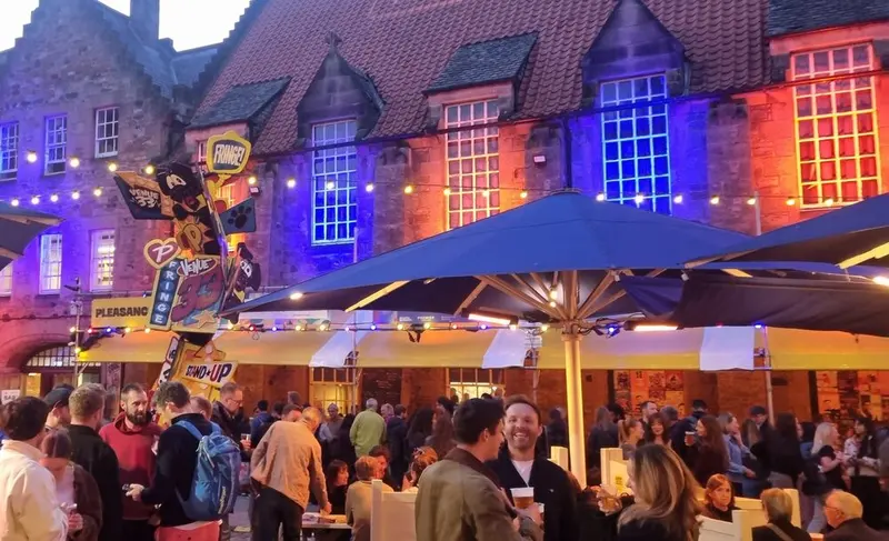 Crowds with drinks in the Pleasance Courtyard at dusk during the 2026 Fringe, under umbrellas, string lights and the venue's