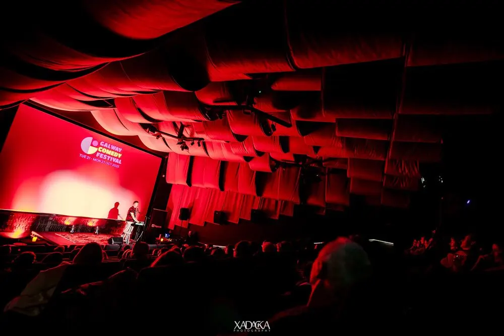 A comedian at the microphone on a red-lit stage in a draped auditorium, the festival's name on the screen behind and the