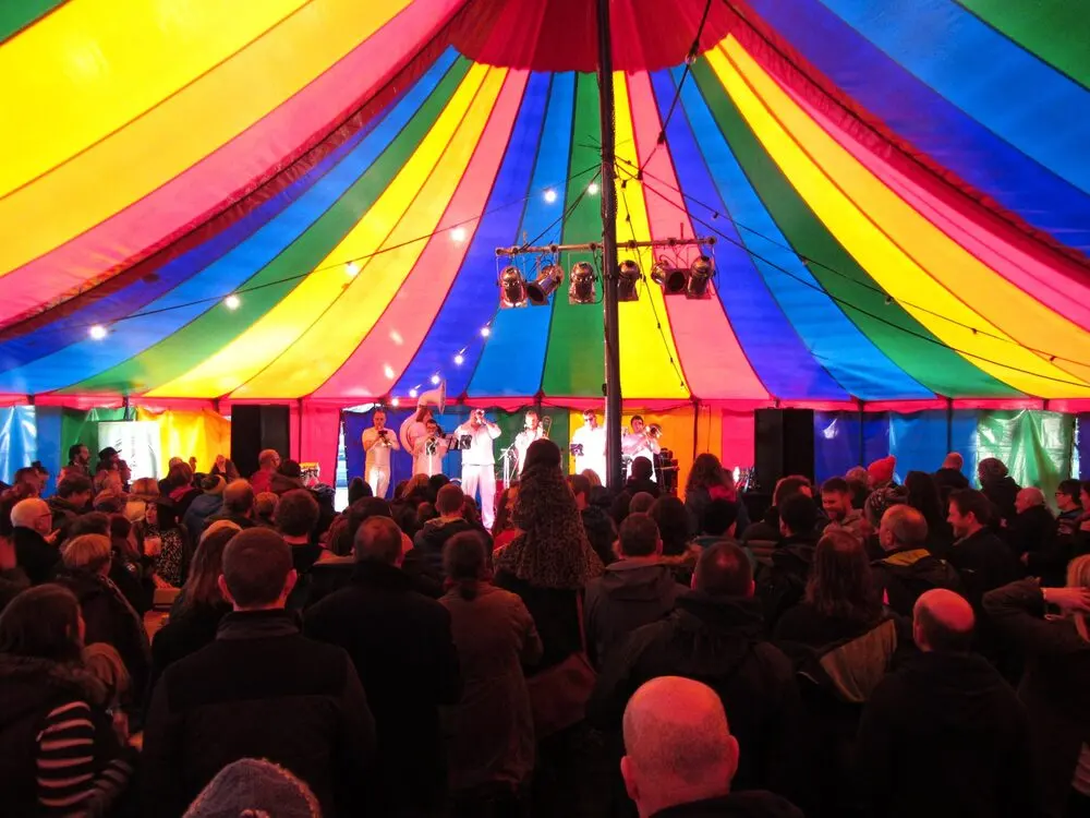 A packed crowd under the striped big top of the festival's music tent, facing a lit stage.