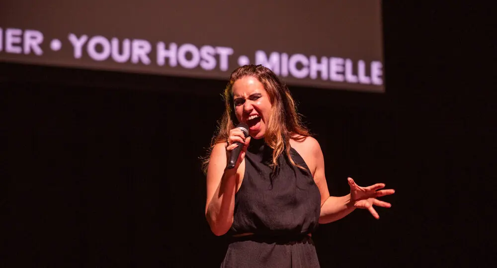 A performer singing into a microphone with arms outstretched on a Melbourne Fringe stage.