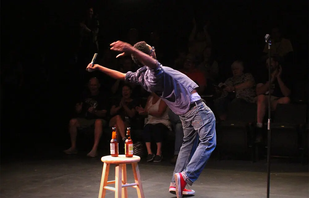 A comedian leaning far back mid-bit on a black-box stage while the front rows laugh and clap