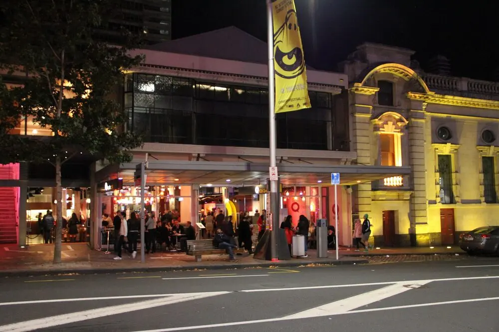 Q Theatre in Auckland at night during the 2015 festival, with a festival banner on the pole and people outside.