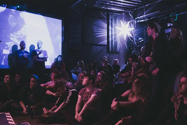 An audience sitting close to the stage under coloured lights at the Black Box, a Cathedral Quarter Arts Festival venue in