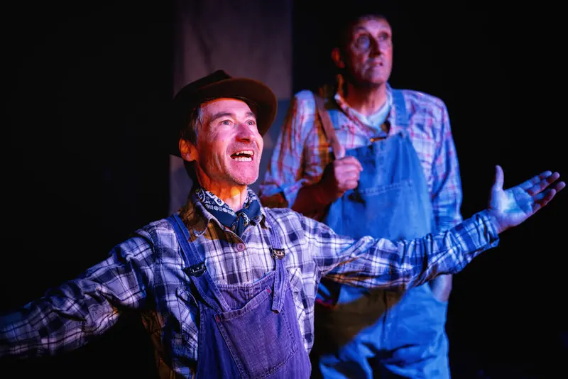 Two performers in overalls and plaid shirts mid-scene under a spotlight at Prague Fringe