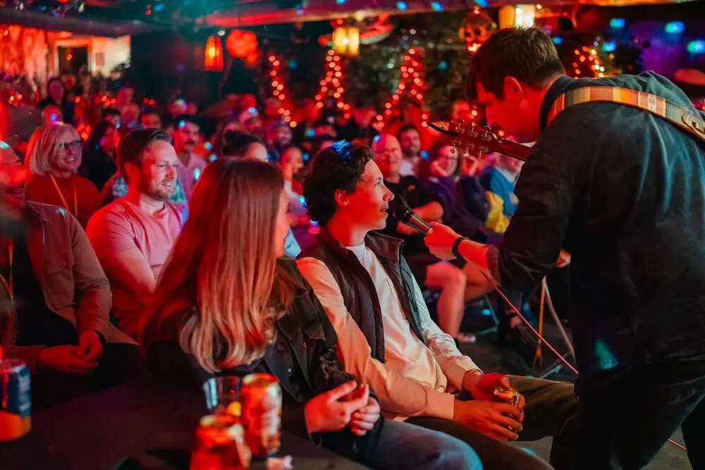 A performer with a guitar playing to the smiling front row of a packed, red-lit room at the Raleigh Comedy Festival