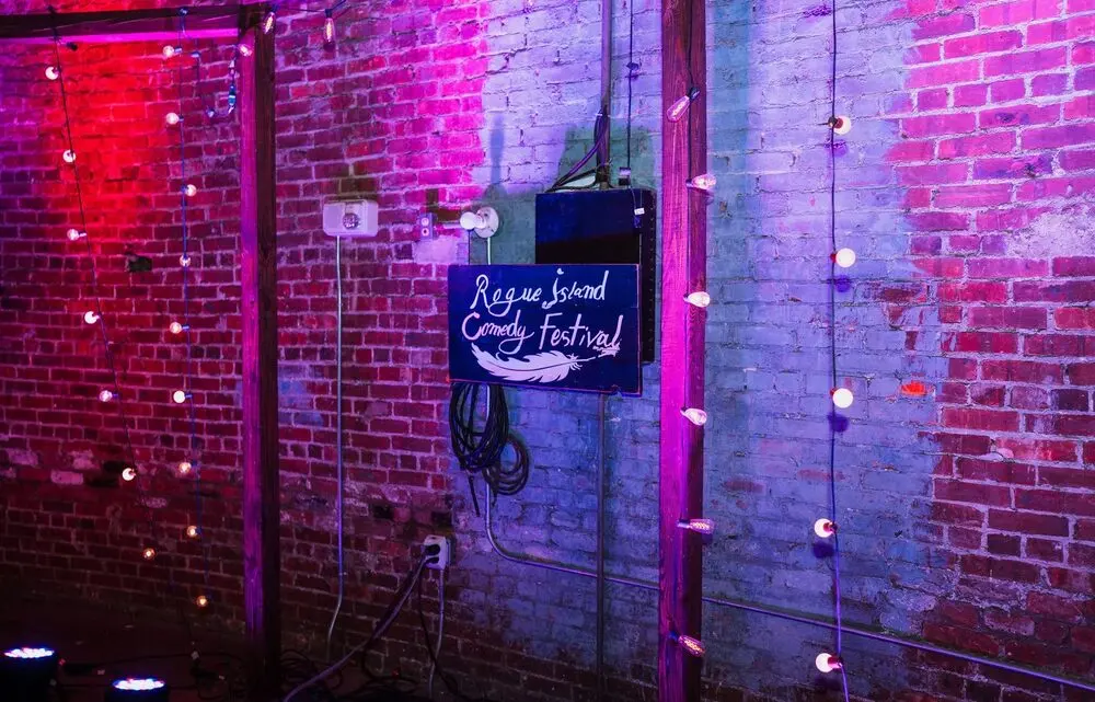 A hand-lettered Rogue Island Comedy Festival sign on a brick wall washed in pink and blue stage light with strings of bulbs