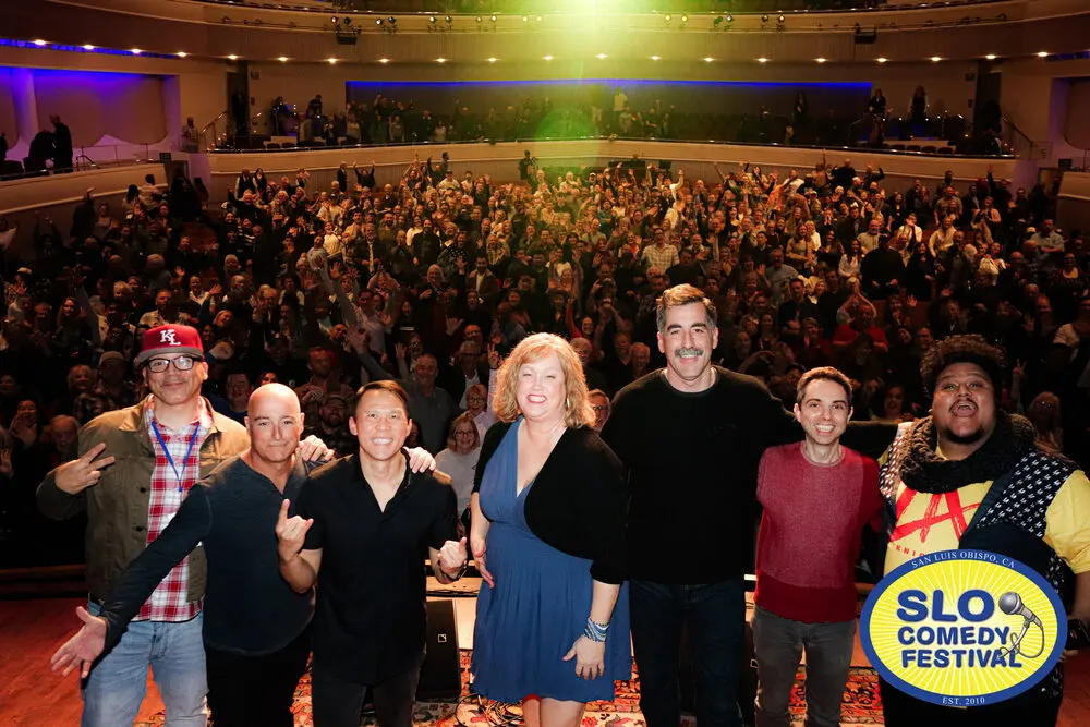 The festival's comedians posing on stage in front of a cheering, full theatre in 2025, with the festival badge in one corner