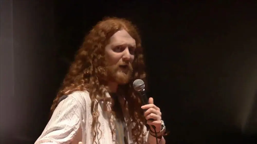 A comedian with long hair performing with a microphone on stage at the Utrecht International Comedy Festival in 2018.