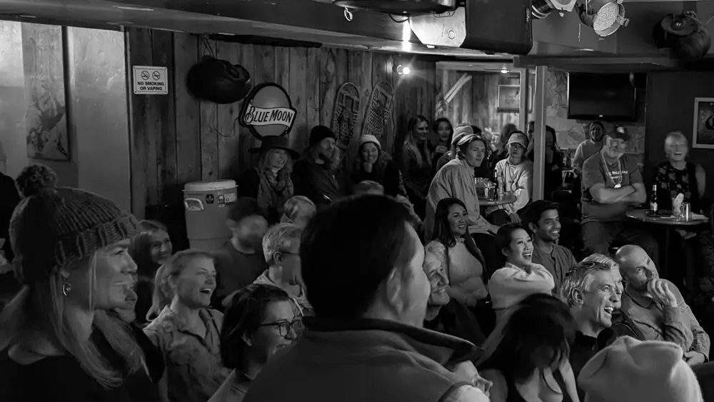 A black-and-white shot of a packed bar audience in winter hats laughing during a Vail Comedy Festival show