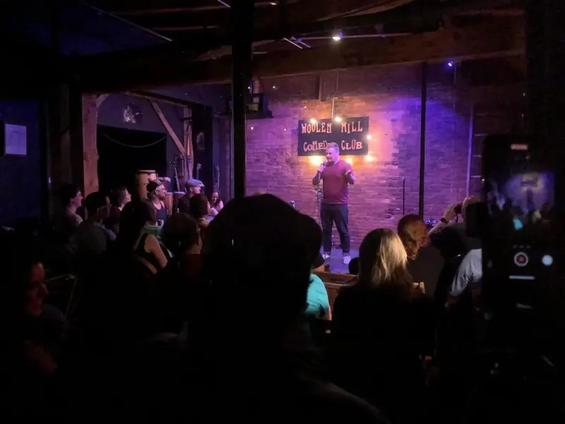 A comedian on stage under the Woolen Mill Comedy Club sign performing to a dark, packed room during the Vermont Comedy