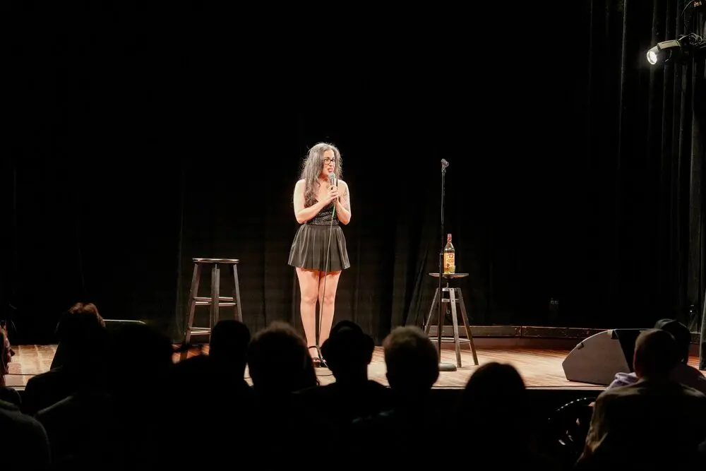 A comedian with a microphone on the Lincoln Hall stage in Chicago, the audience in silhouette, August 2026
