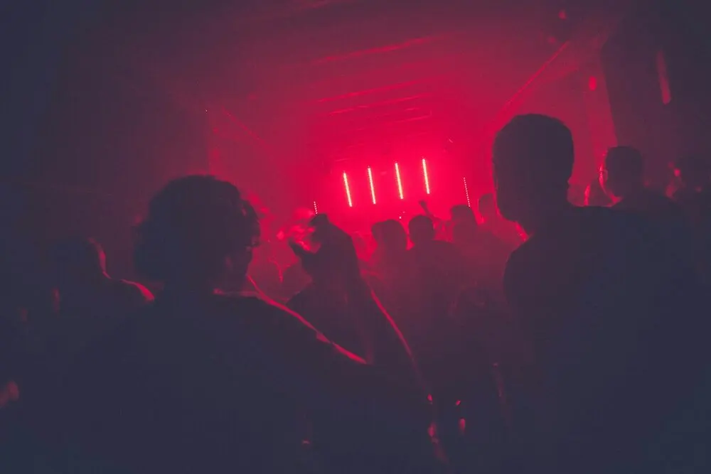 A packed small room seen from the back, heads in silhouette, red stage light ahead