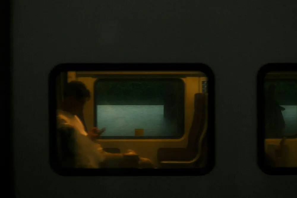 A lit train window at night, a blurred passenger looking down at a phone