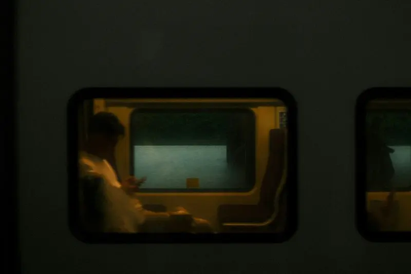 A lit train window at night, a blurred passenger looking down at a phone