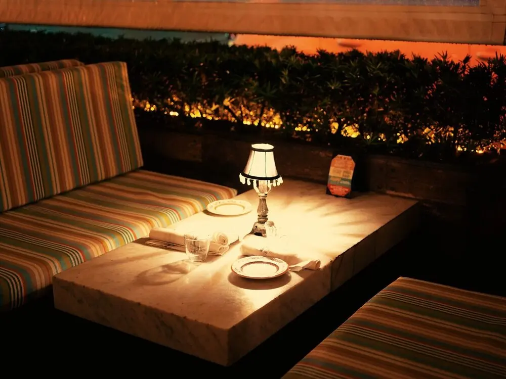 A late-night booth: one lamp on a table set with plates and glasses, striped benches, fairy lights in the hedge