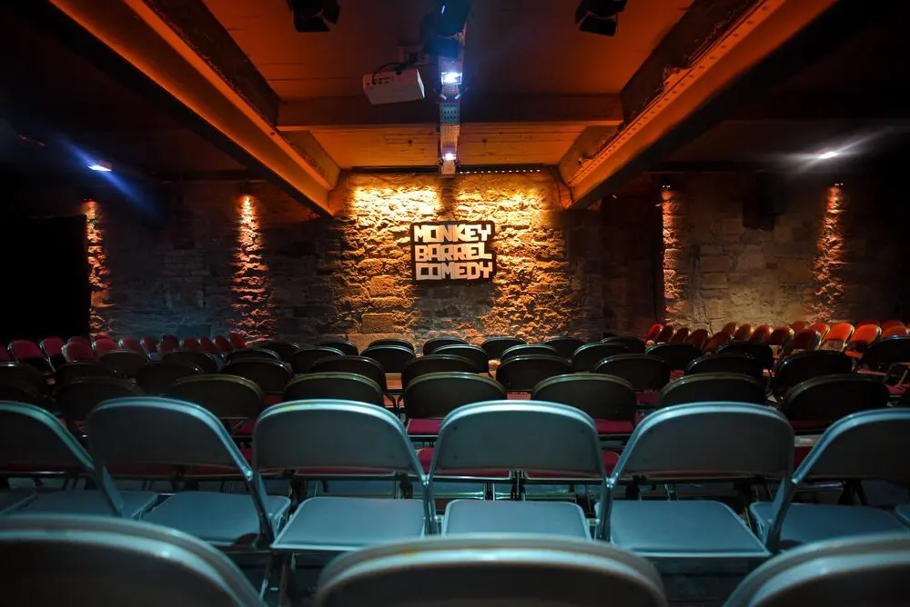 The empty showroom: rows of chairs facing a small stage, the Monkey Barrel Comedy sign lit on a stone wall in warm light.