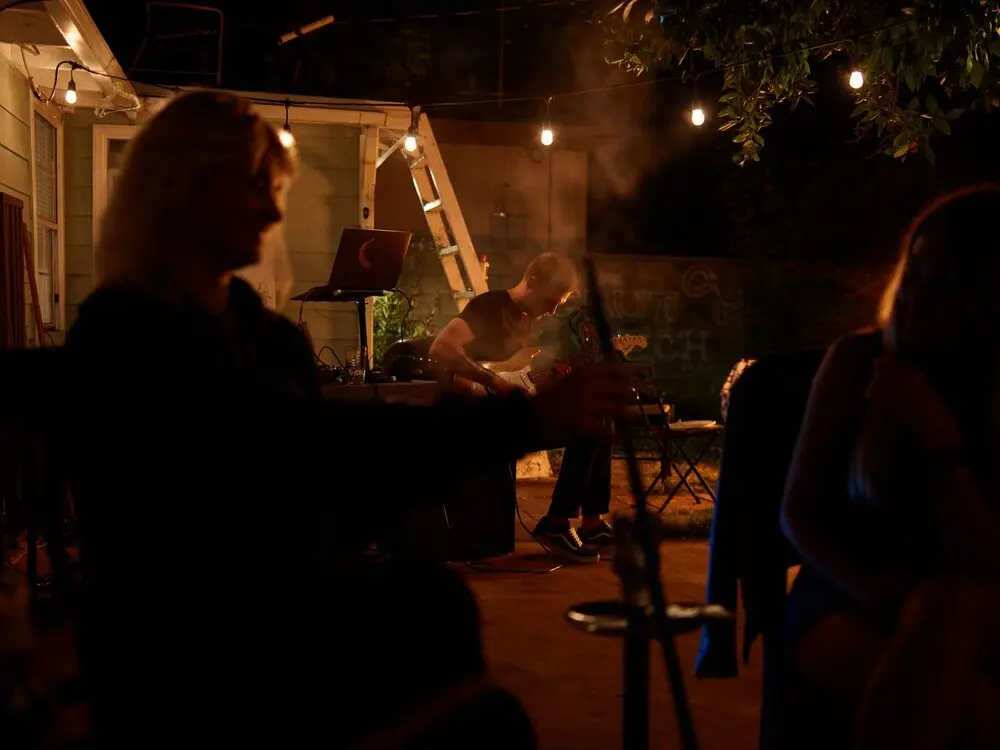 Backyard show at night: string lights, folding chair, guitarist, small audience; faint painted letters on back wall