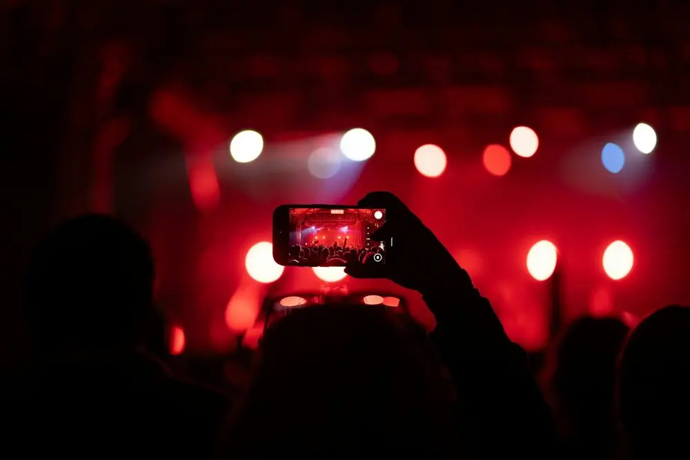 A phone held up over a silhouetted crowd, filming a red-lit stage